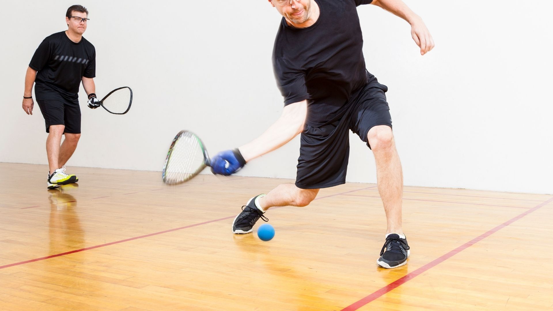 Man swinging racquet at racquetball while another man prepares to play Man swinging racquet at racquetball while another man prepares to play