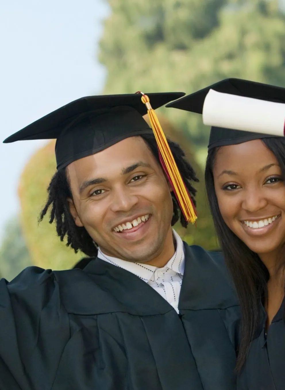 2 smiling young people in graduation gowns and caps holding diplomas