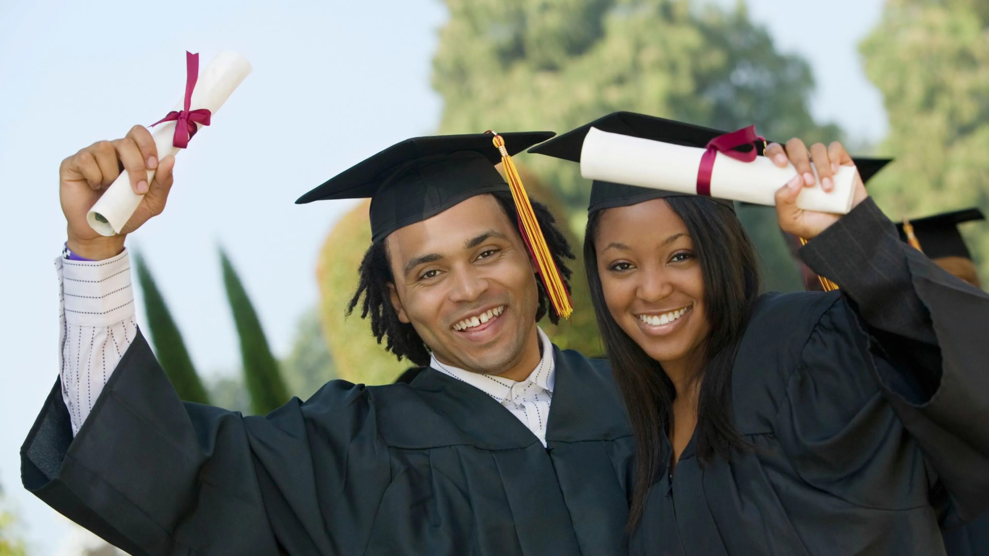 2 smiling young people in graduation gowns and caps holding diplomas